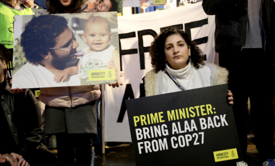 Image of Sanaa, Alaa's sister, holding a placard at a protest that reads: Prime Minister Bring Alaa back from COP27. Beside her someone is holding an image of Alaa and his baby son. 