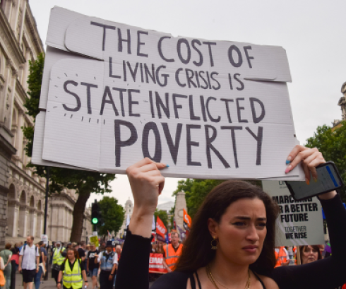 Person holding a sign up in a protest which reads "The Cost of Living Crisis is State Inflicted Poverty"